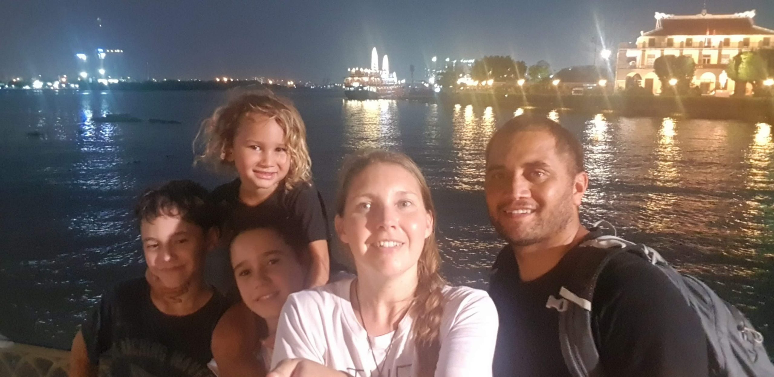 Family photo taken with a selfie stick along the riverside in Saigon with city buildings and water in the background