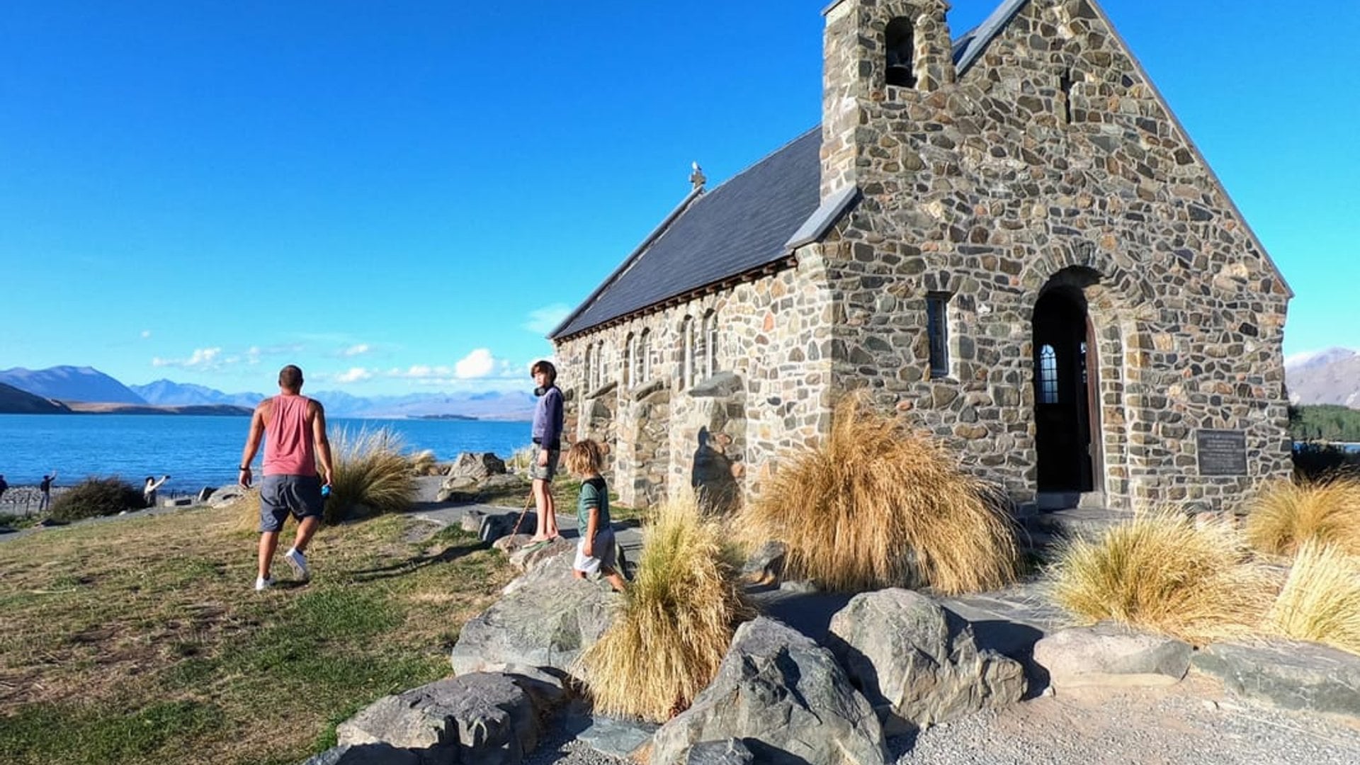 Family exploring the iconic Church of the Good Shepherd at Lake Tekapo with turquoise waters and snow-capped mountains in New Zealand